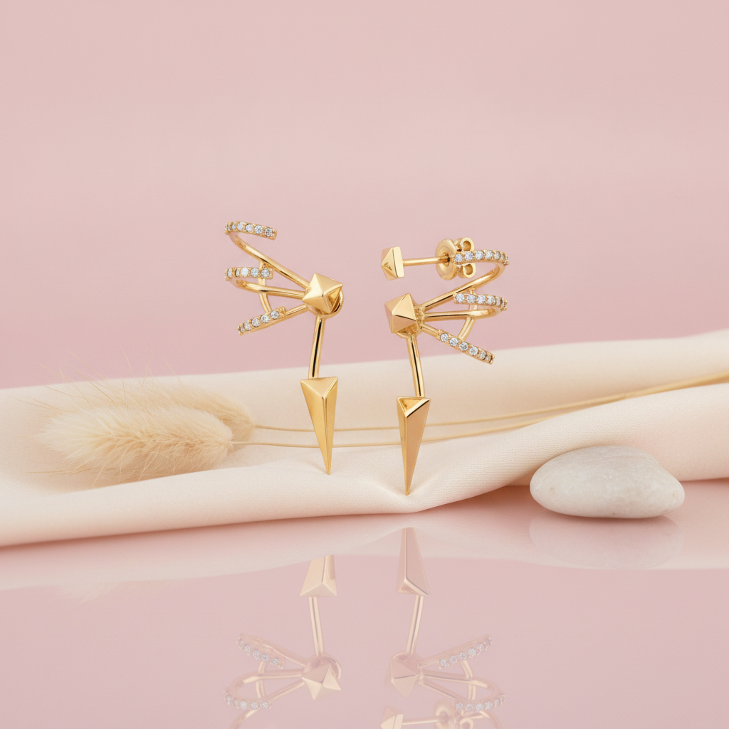 gold sculptural earrings with curved pave bands, a faceted pyramid center and a sharp spike, styled with dried grasses on a pink background