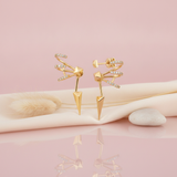gold sculptural earrings with curved pave bands, a faceted pyramid center and a sharp spike, styled with dried grasses on a pink background