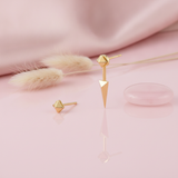gold faceted pyramid stud earring and a pyramid-topped spike drop stud earring on a pink background with dried grass