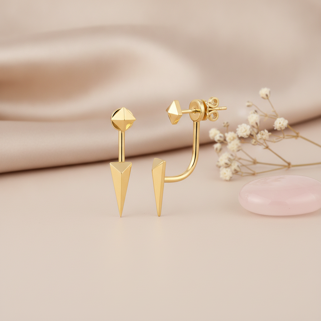 gold pyramid stud and spike jacket earrings styled on a blush fabric backdrop with dried flowers and a pink stone