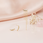 mismatched gold curved bar earrings with pave stones, one side trailing cascading chains, styled on a blush fabric backdrop with dried flowers