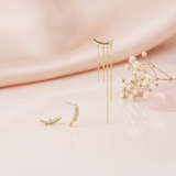 mismatched gold curved bar earrings with pave stones, one side trailing cascading chains, styled on a blush fabric backdrop with dried flowers