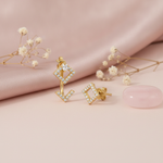 gold rhombus stud earrings with a pear-cut stone and floating chevron jacket, styled on a blush fabric backdrop with dried flowers and a pink stone