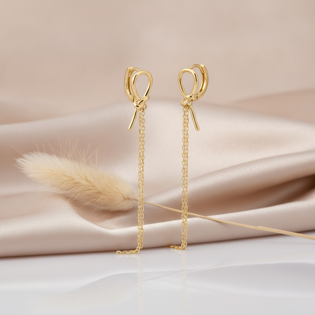 gold knot huggie hoop earrings with trailing drop chains styled on a blush fabric backdrop with a dried flower stem