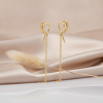 gold knot huggie hoop earrings with trailing drop chains styled on a blush fabric backdrop with a dried flower stem