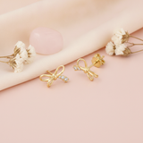 gold bow stud earrings with open loops and accent stones styled on a blush fabric backdrop with dried flowers and a pink stone