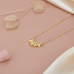 gold sunburst pendant necklace styled with dried leaves and a pink stone on a blush background