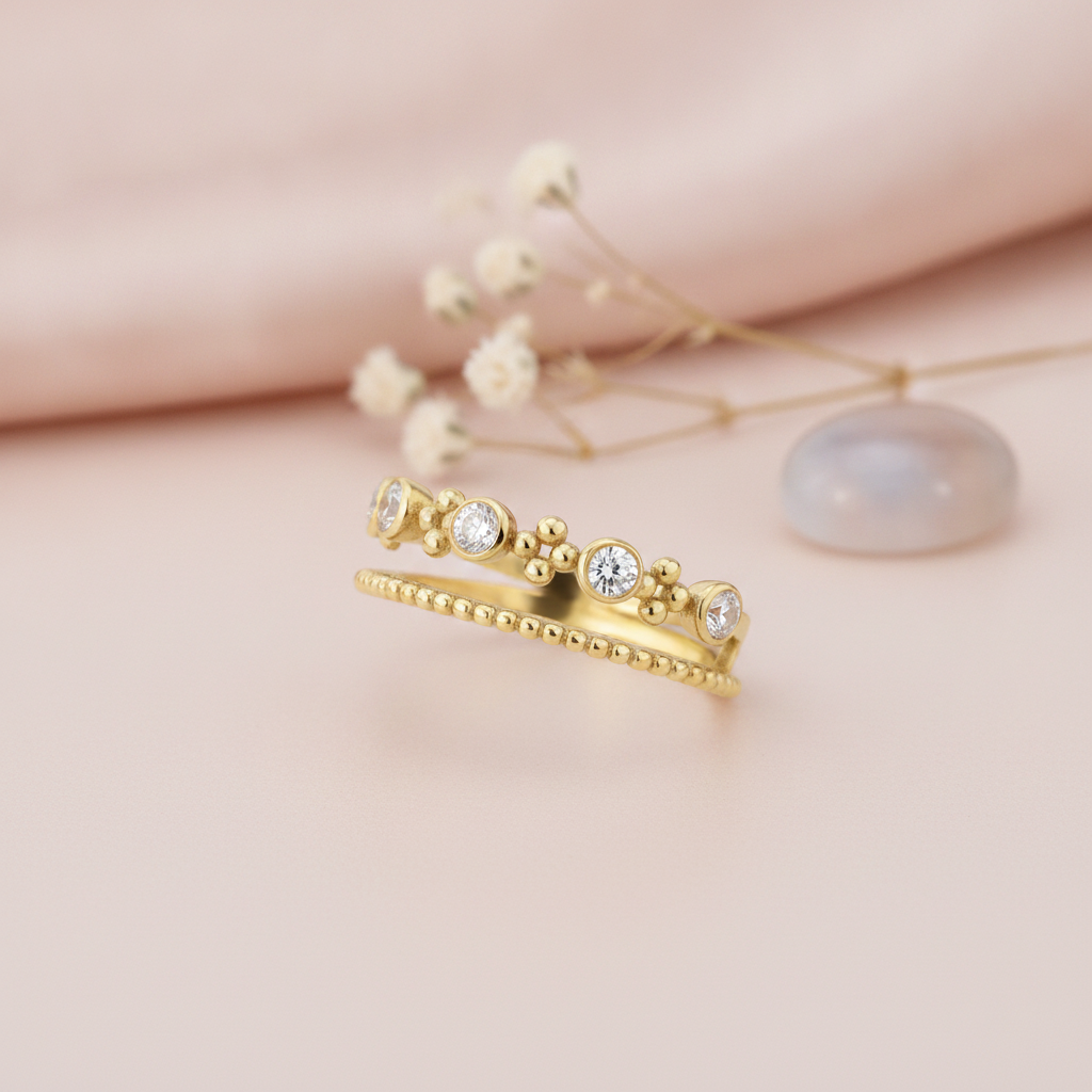 double-band gold ring with a beaded strand, bezel-set stones, and polished gold spheres, styled with baby's breath and a gray-blue stone on a pink background