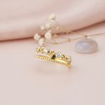 double-band gold ring with a beaded strand, bezel-set stones, and polished gold spheres, styled with baby's breath and a gray-blue stone on a pink background