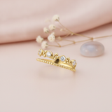 double-band gold ring with a beaded strand, bezel-set stones, and polished gold spheres, styled with baby's breath and a gray-blue stone on a pink background