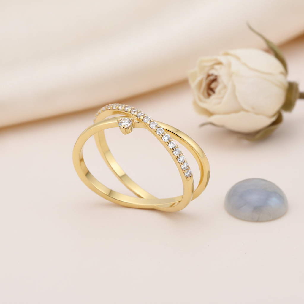 crossover gold ring with a paved band and a single suspended clear stone, styled with a white rose and a gray-blue stone on a cream background