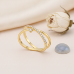 crossover gold ring with a paved band and a single suspended clear stone, styled with a white rose and a gray-blue stone on a cream background