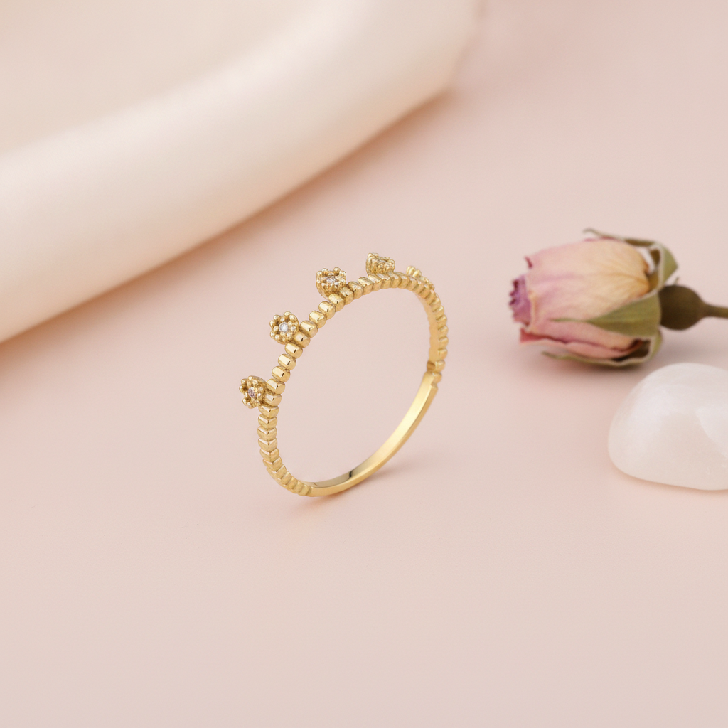 gold ring with floral crests set with clear stones on a beaded band, styled with a dried rose on a blush background