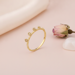 gold ring with floral crests set with clear stones on a beaded band, styled with a dried rose on a blush background