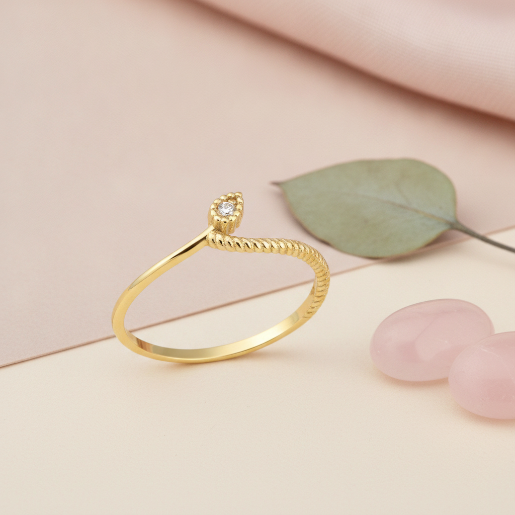gold bypass ring with a twisted rope profile and a beaded drop stone setting, styled with a dried leaf and pink stones on a blush background