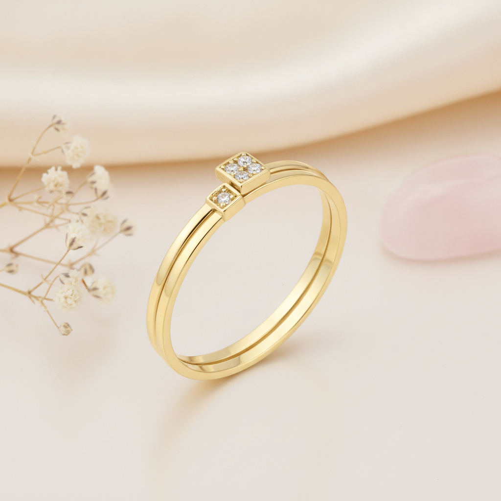 gold ring with two offset square settings of clear stones, on cream fabric with dried flowers and a pink stone