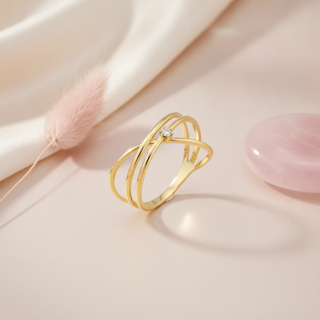 gold ring with crossing architectural bands and a single small clear stone, on blush fabric with a pink feather prop