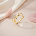 gold ring with crossing architectural bands and a single small clear stone, on blush fabric with a pink feather prop