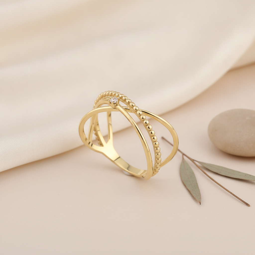 gold crossover ring with a beaded strand and smooth arcs, on blush fabric with a dried grass stem