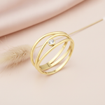 wide open gold ring with overlapping arcs and a single bezel-set clear stone, on cream fabric