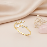 asymmetric gold bypass ring with a cluster of pear, marquise, and round clear stones, on cream fabric with a pinecone flower