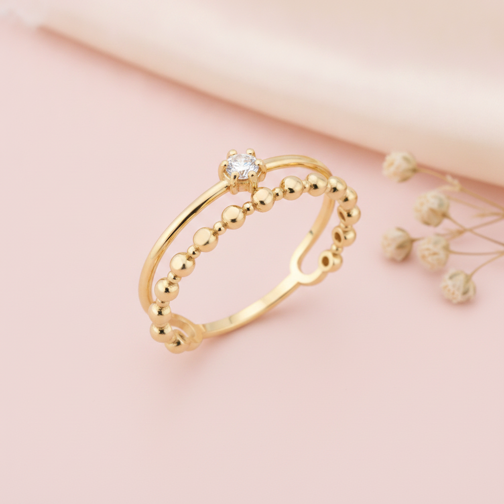 double gold ring with a smooth arch, a single clear stone, and a beaded band, on pink fabric with dried flowers
