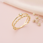 double gold ring with a smooth arch, a single clear stone, and a beaded band, on pink fabric with dried flowers