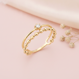 double gold ring with a smooth arch, a single clear stone, and a beaded band, on pink fabric with dried flowers