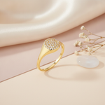 gold signet ring with a flat circular crest paved in clear stones, on cream fabric with a dried flower