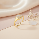 gold signet ring with a flat circular crest paved in clear stones, on cream fabric with a dried flower