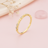 gold ring with raised vertical bar accents and small square-set clear stones on blush silk with dried flowers and rose quartz