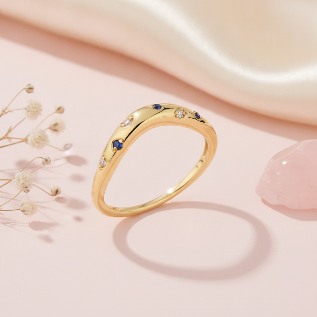 gold ring with scattered blue and clear stones on a smooth wave-shaped band on blush silk with dried flowers and rose quartz