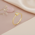 gold ring with a clear stone set in a square frame, styled with dried baby's breath, a stone, and dried grass on a blush background