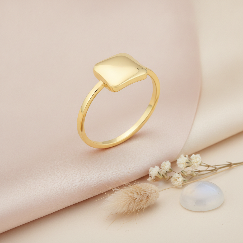 gold ring with a cushioned square set diagonally, styled with dried baby's breath, a stone, and dried grass on a blush background