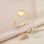 gold ring with a cushioned square set diagonally, styled with dried baby's breath, a stone, and dried grass on a blush background