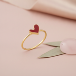 gold Cora heart ring with crimson enamel on a blush pink background with a rose quartz bead and leaf accent