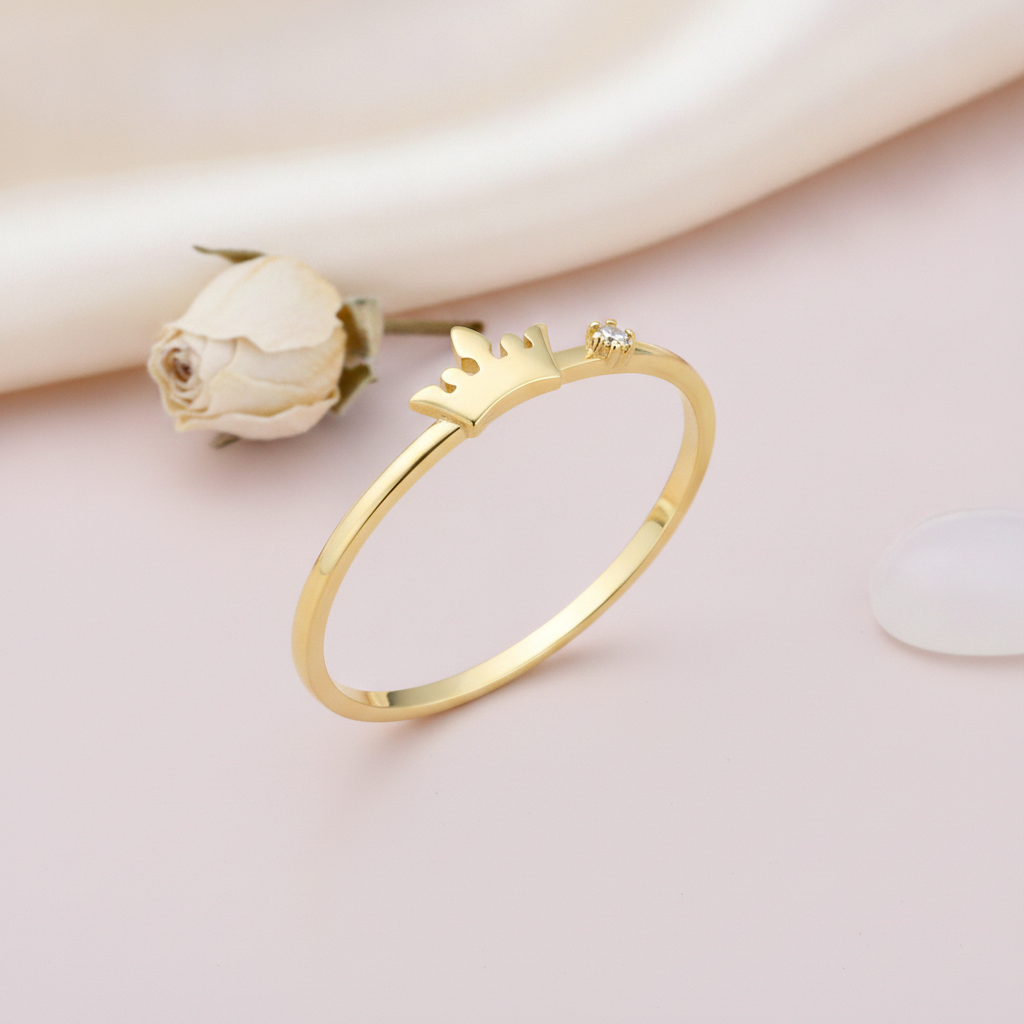 gold crown motif ring with a single clear stone on a blush background with a dried rose and a white stone
