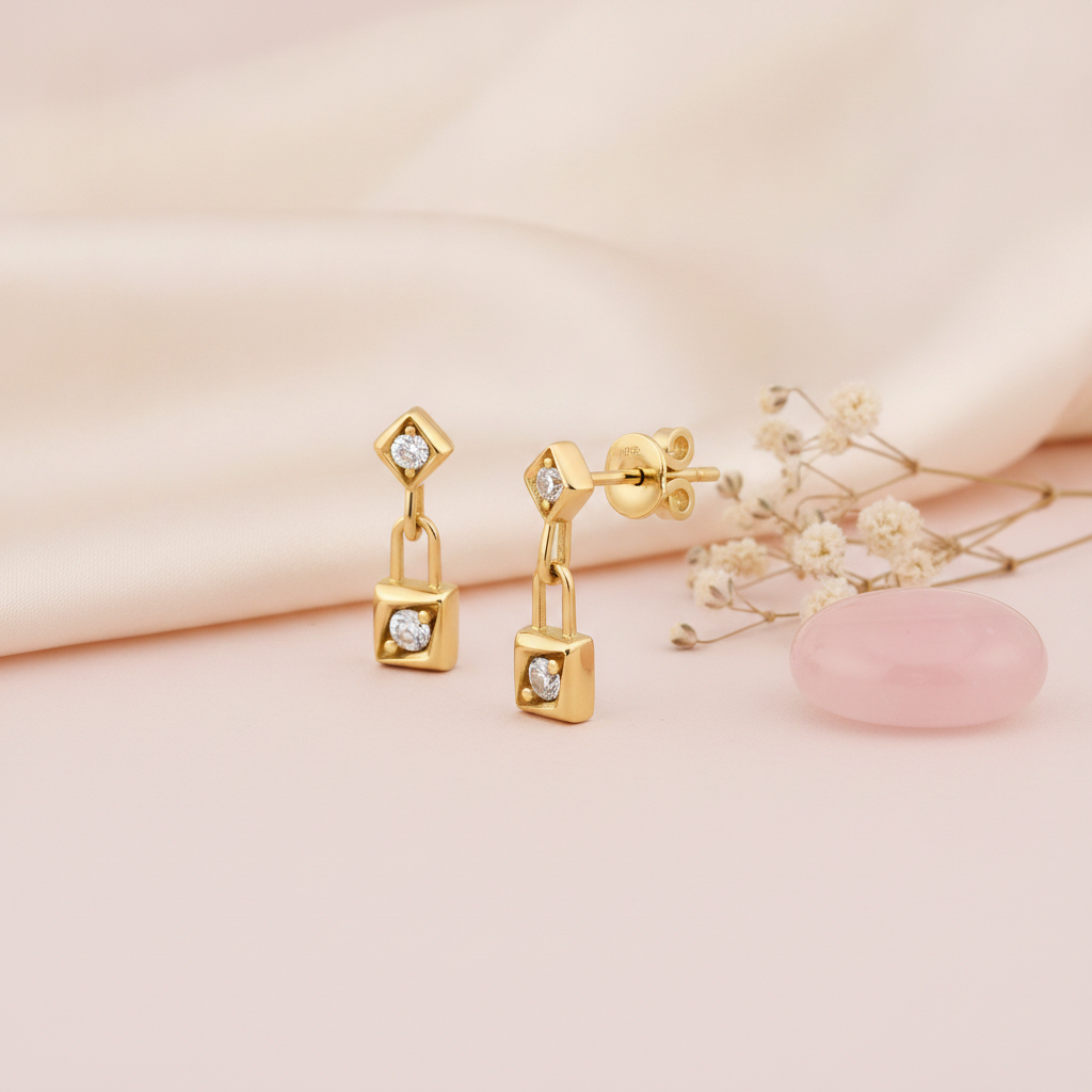 gold kite-shaped drop earrings with a linked square charm and clear stones on a cream background with baby's breath and a pink stone