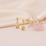 gold kite-shaped drop earrings with a linked square charm and clear stones on a cream background with baby's breath and a pink stone