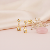 gold kite-shaped drop earrings with a linked square charm and clear stones on a cream background with baby's breath and a pink stone