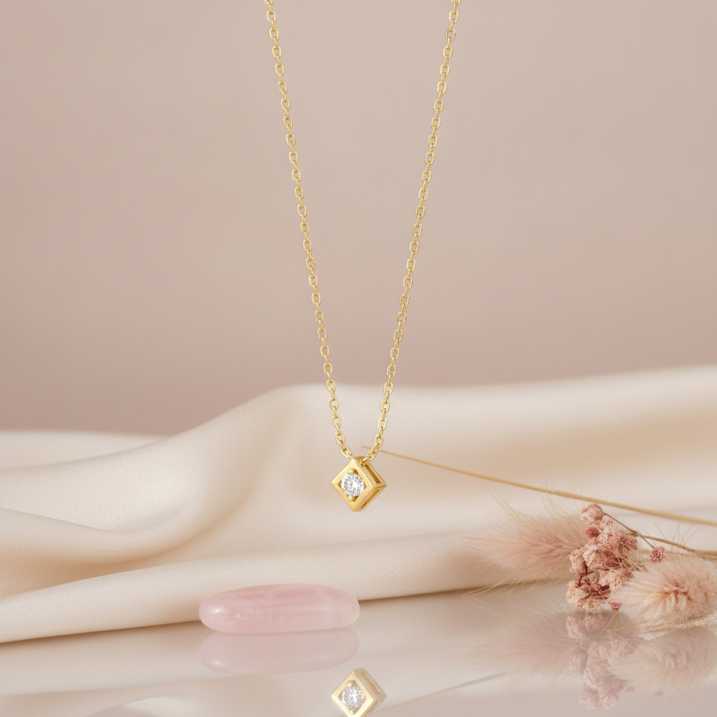 gold tilted square pendant necklace with a clear stone on a mauve background with draped satin fabric and pink pampas grass