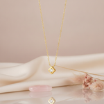 gold tilted square pendant necklace with a clear stone on a mauve background with draped satin fabric and pink pampas grass