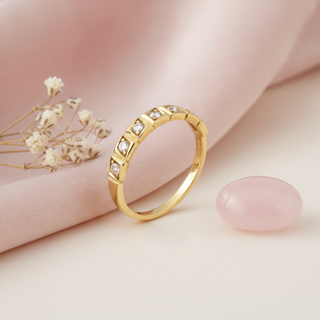 gold ring with a continuous row of raised square settings and clear stones on a pink background with baby's breath and a rose quartz stone