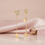 asymmetrical gold star drop earrings with White CZ stones and dangling star charms on a blush background with a dried rose and a pink stone