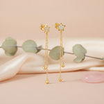 gold star stud earrings with cascading chains and polished spheres on a blush pink backdrop with eucalyptus leaves and a rose quartz stone