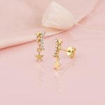 gold stud earrings with a stone-set star arc and a dangling star charm on a pink backdrop with a dried flower