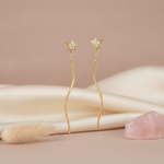 gold star stud earrings with slender wavy drop chains on a pink backdrop with dried grass and a rose quartz stone