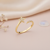 gold open ring with a stone-set star and a curved trailing arc on a pink backdrop with dried flowers and a moonstone