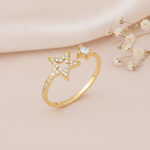 gold open bypass ring with a stone-set star outline and a princess-cut stone on a pink backdrop with dried baby's breath