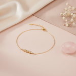 gold chain bracelet with a curved cluster of marquise and round stones on a pink backdrop with a logo tag charm, dried flowers, and a rose quartz stone